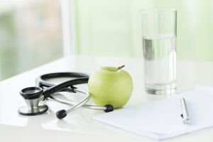 Healthy lifestyle items: green apple, glass of water, stethoscope and blank notebook on the table.