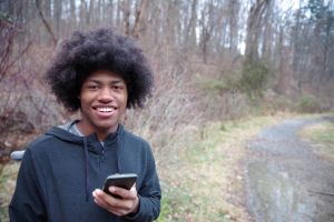 African American Teenager Smiles Using His Mobile Phone On A Wooded Hiking Trail