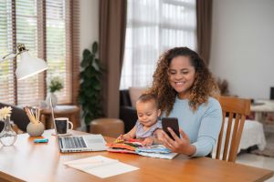 Mother at home with baby working on laptop, holding smartphone