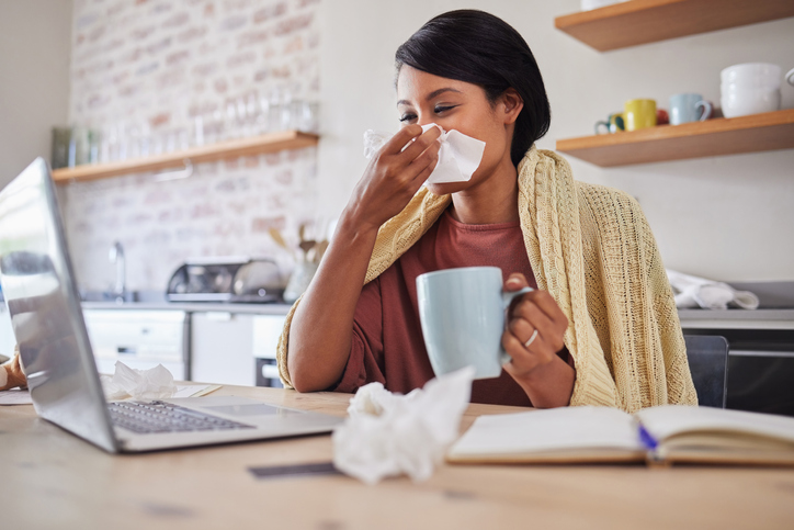 Sick, tea and business woman with covid while reading an email on a laptop and working from house. Remote entrepreneur blowing nose with flu and drink of coffee while planning work on web in her home