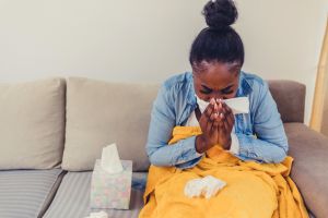 Sick young woman sitting on sofa blowing her nose at home in the sitting room. Photo of sneezing woman in paper tissue.