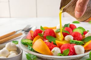 Pouring dressing in fruit summer salad made of fresh fruits, mozzarella cheese, cucumbers and mint on white table