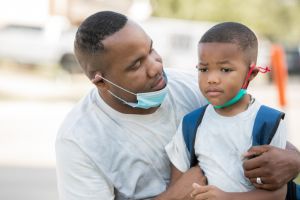 Father consoles anxious kindergartener while they wear masks as he's dropping him off for first day of school,