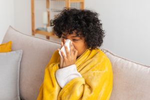 Unhappy sick young african american woman with cold sitting on sofa at home. Girl covering with blanket and blowing her nose in tissue. Medical and health concept. Cold flu allergy. Sick at home.