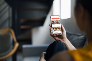 Over the shoulder view of young woman ordering food online from the menu via mobile app on smartphone, lying on sofa in the living room at home. Online food delivery and dining mobile app. Lifestyle and technology