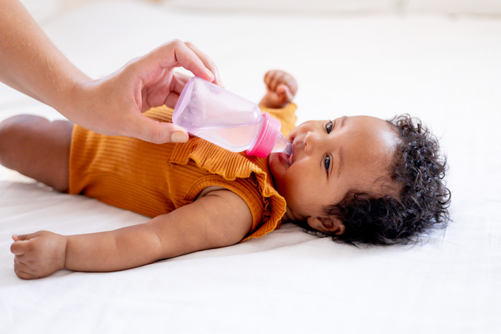 a small African-American baby girl drinks water or milk from a bottle in an orange bodysuit on a white bed, her mother's hand feeds a six-month-old black newborn baby from a bottle