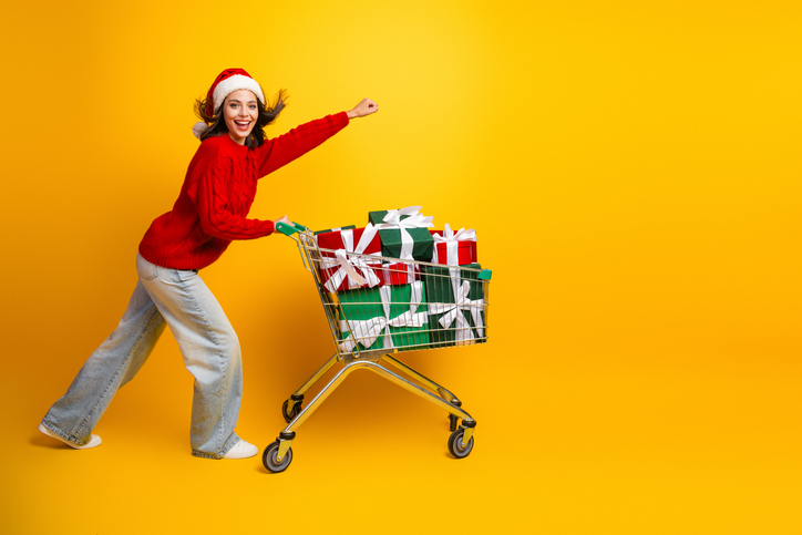 Cheerful santa hat wearing shopper pushes a cart loaded with gifts through a bright yellow studio smiling and laughing ready for Christmas shopping and holiday fun