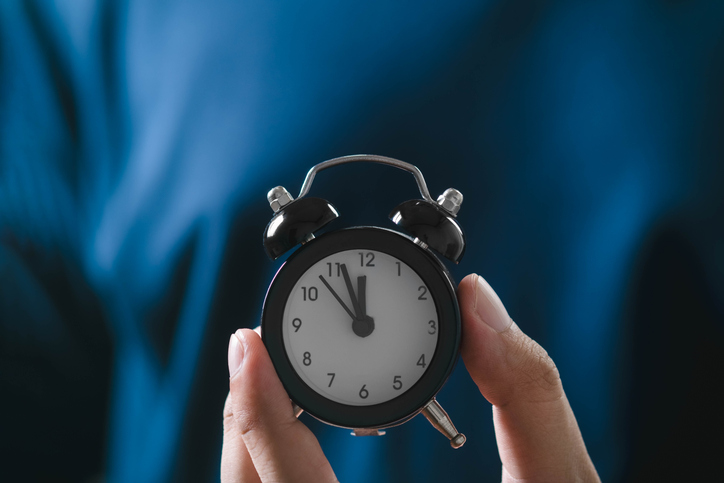 Close-up of hands holding a small black alarm clock in front of a dark blue background, symbolizing time management and punctuality.
