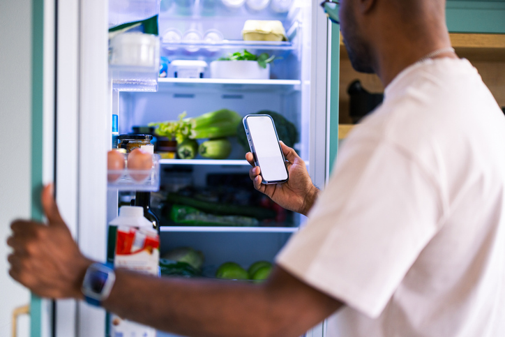 Man With Phone By Open Fridge Full Of Fresh Produce