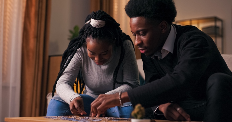 Two siblings sit together working on a puzzle, focused on completing the image. The sister has long hair, and the brother wears a black suit. They chat and enjoy each other's company.