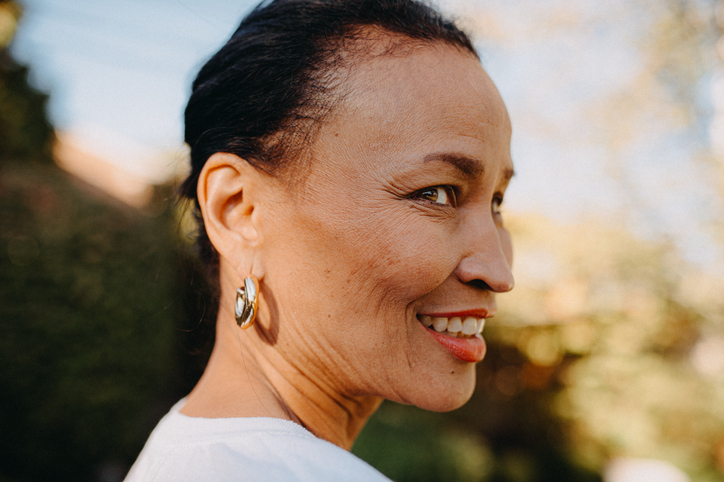 A senior woman is shown in a profile view, smiling softly under warm sunlight.