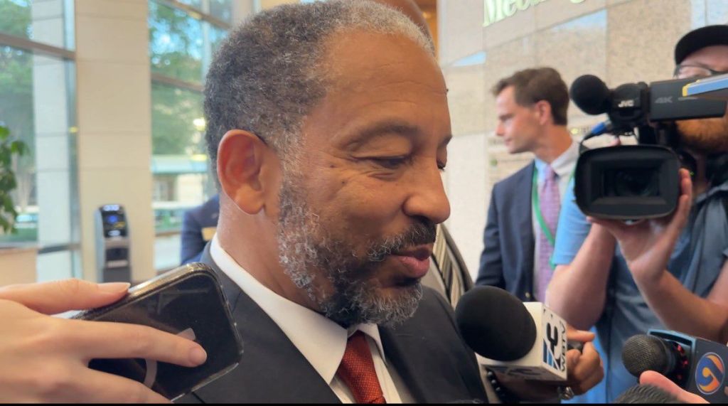 A man with a gray beard and suit being interviewed by multiple reporters holding microphones.
