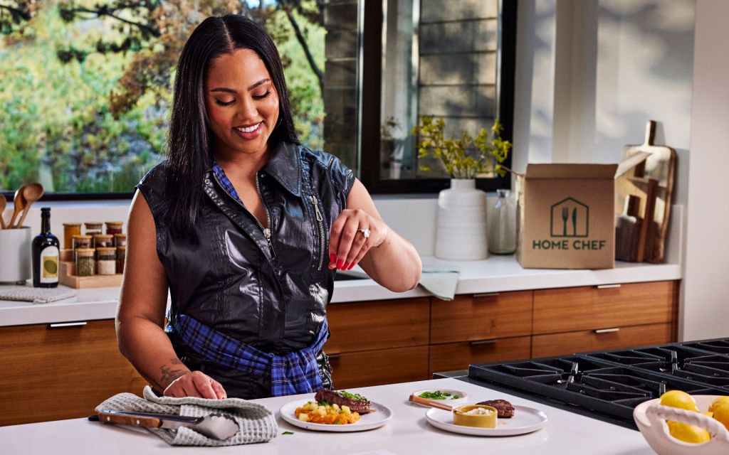 A smiling woman in a black leather vest stands in a kitchen, preparing a meal on a wooden countertop with a "Home Chef" box visible.
