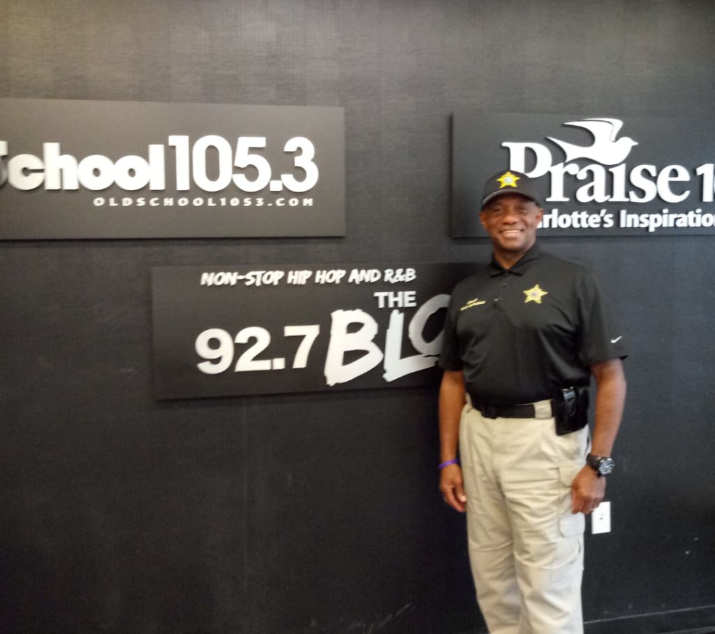 A smiling man in a black uniform stands in front of signs for radio station WBCN 92.7 and Oldschool 105.3.