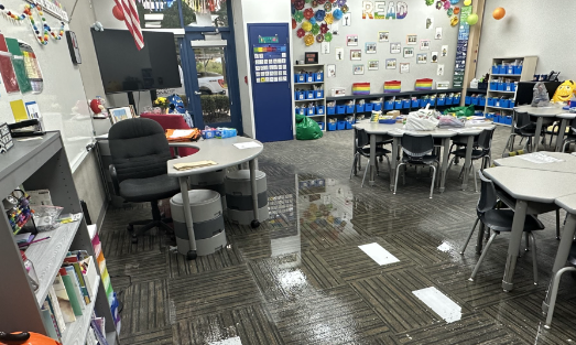 Classroom with desks, chairs, and shelves displaying educational materials and supplies.