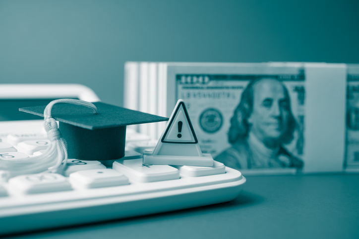 Warning sign and mortarboard on white calculator and heap US dollar banknotes.
